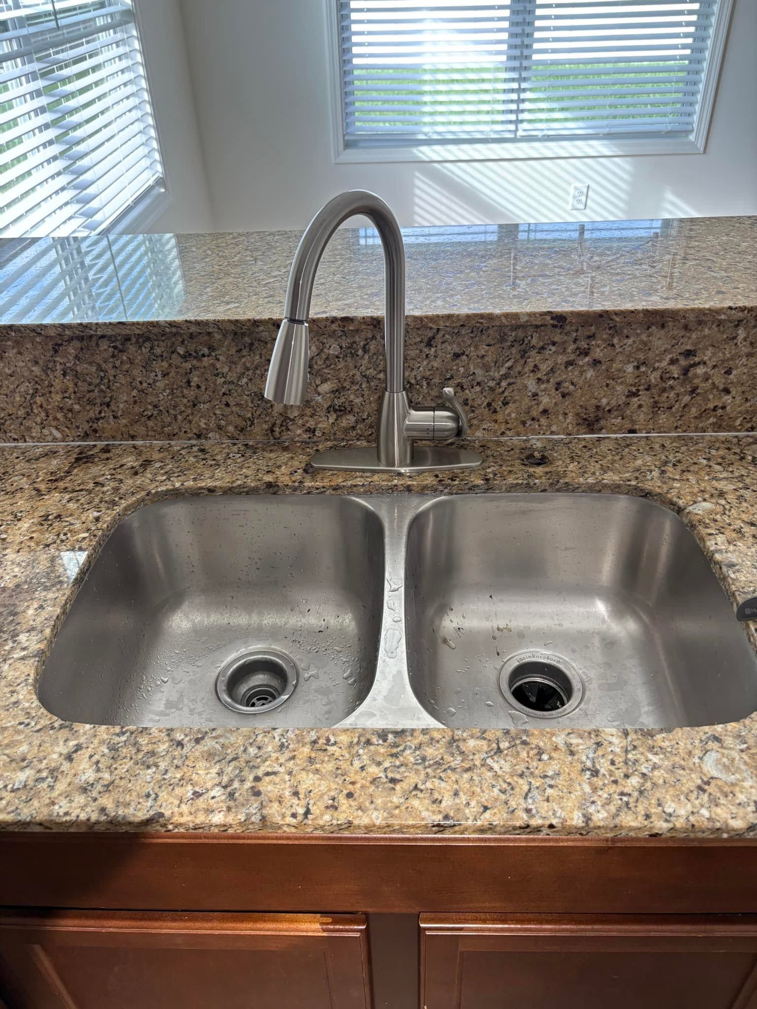 Double kitchen sink under a window, with granite countertop and chrome faucet.