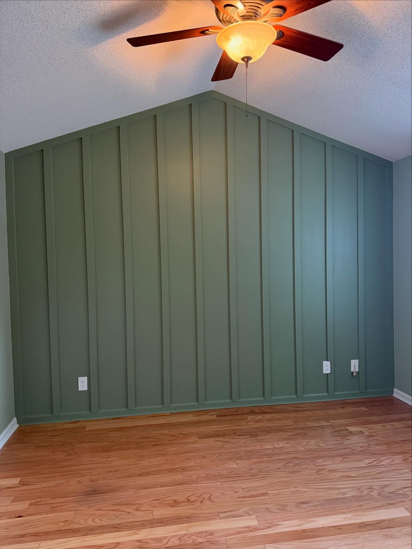Empty room with green paneled accent wall, hardwood floor, and ceiling fan lit overhead