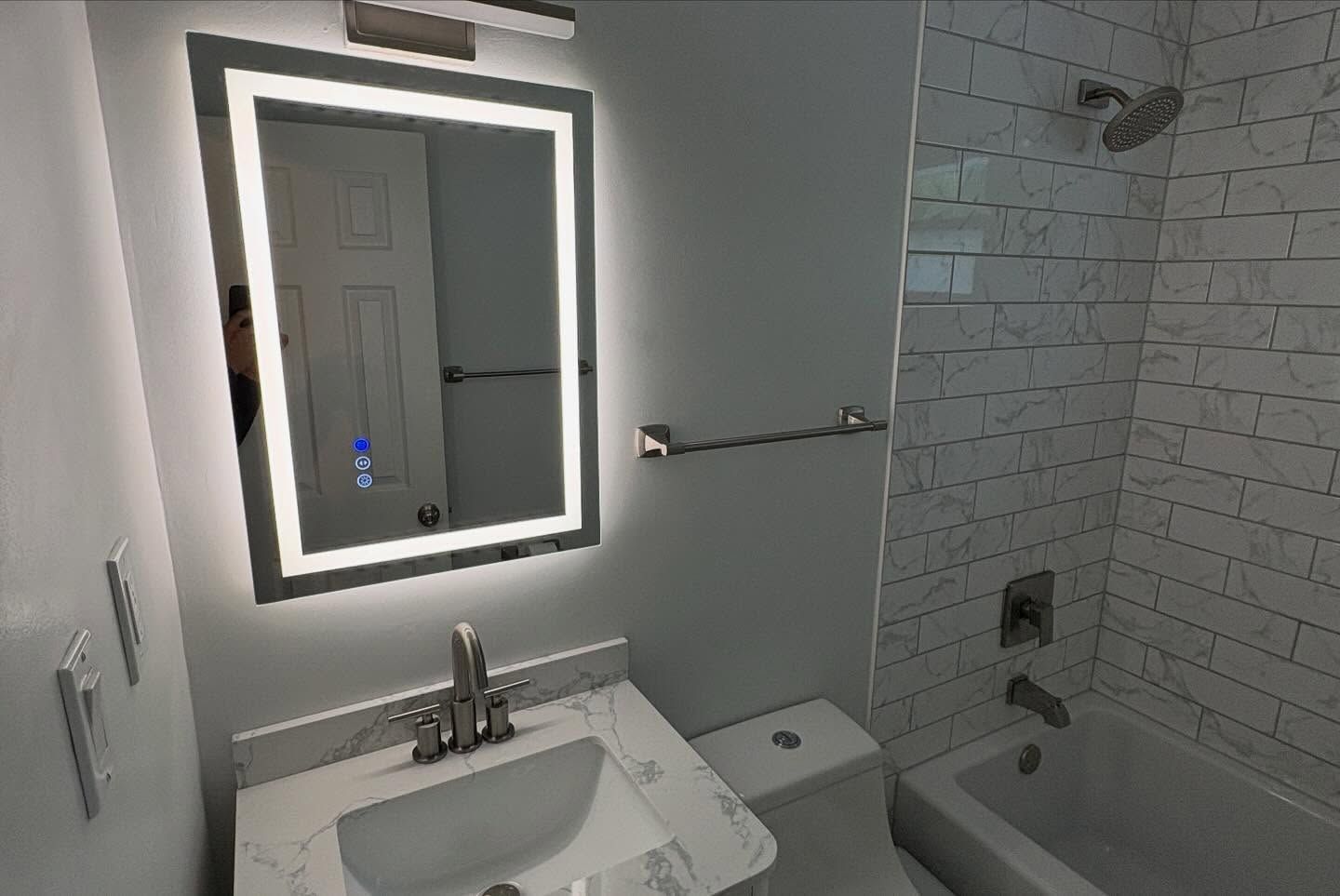 Modern bathroom with lit mirror, white vanity, toilet, towel bar, and subway-tile shower