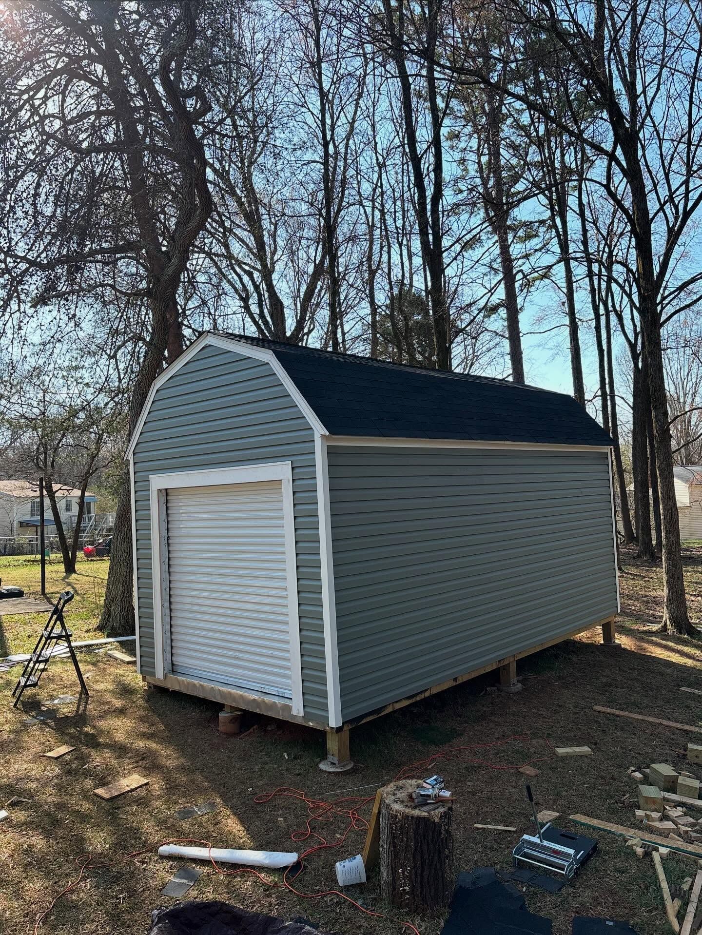 Small gray storage shed with white trim and roll-up door, outdoors among bare trees.