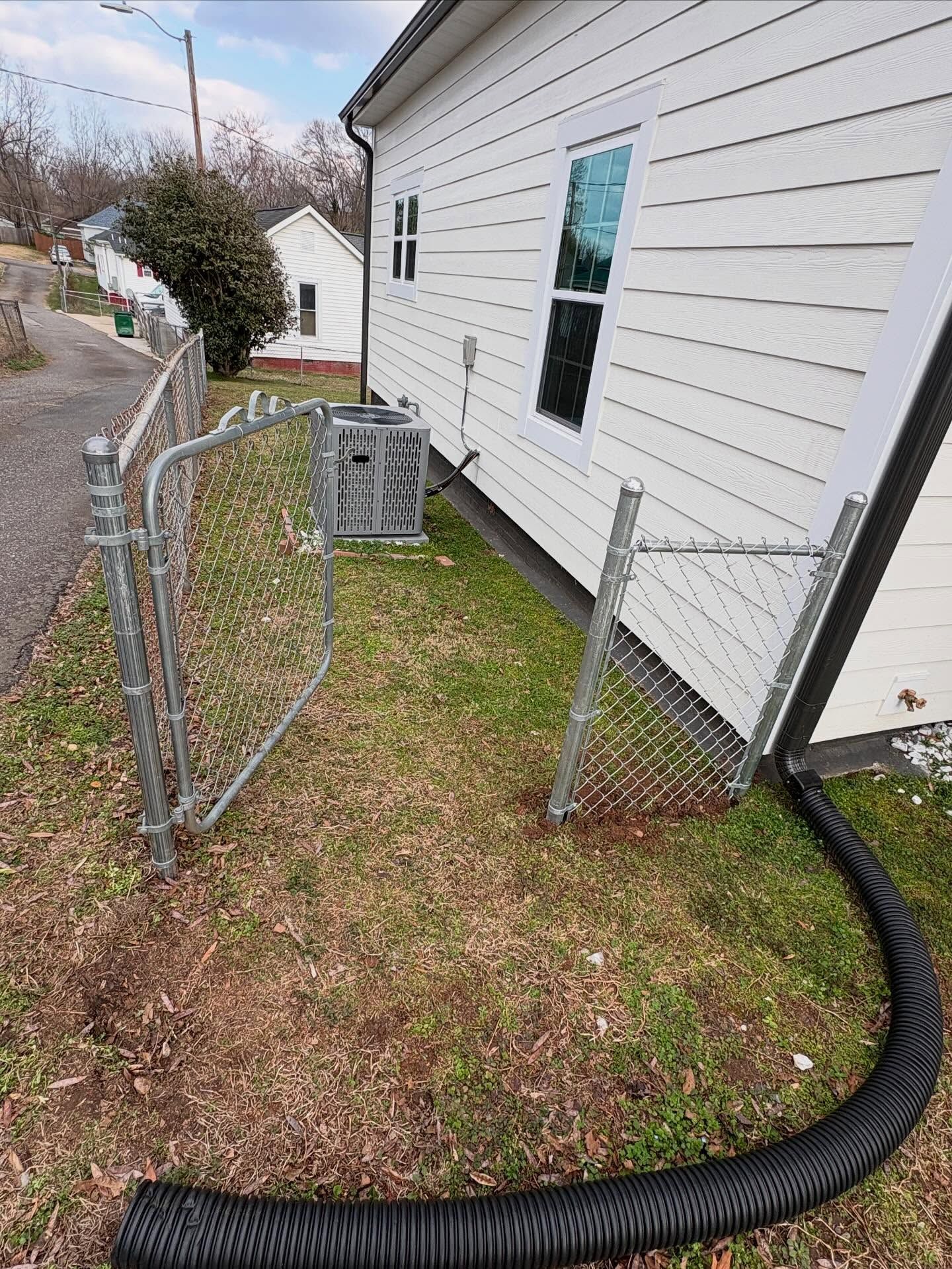 Open chain-link gate beside a white house, with a black drainage hose across the grass.