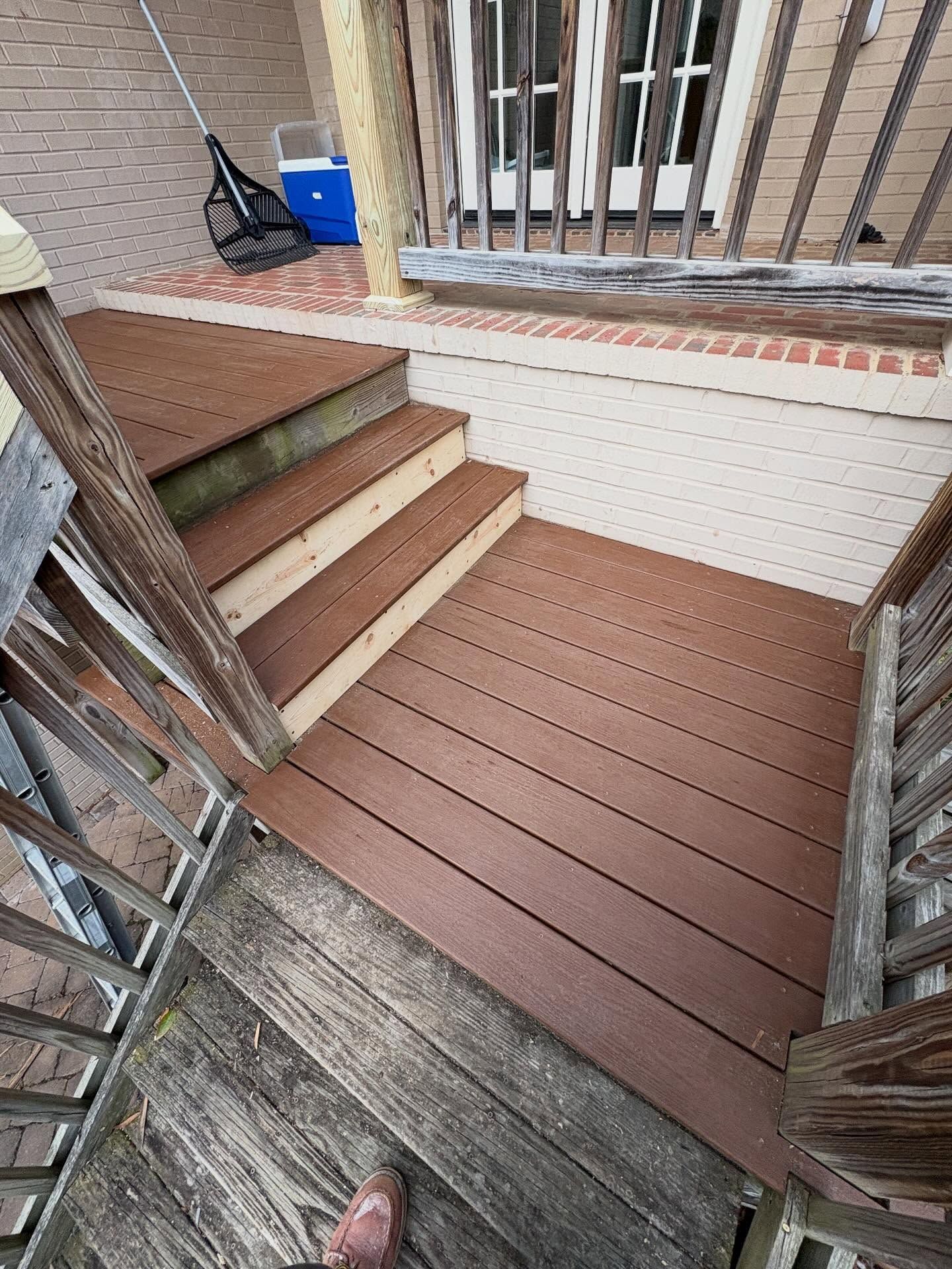 Small outdoor deck with brown stairs, white railing, and a blue bucket near a brick wall.