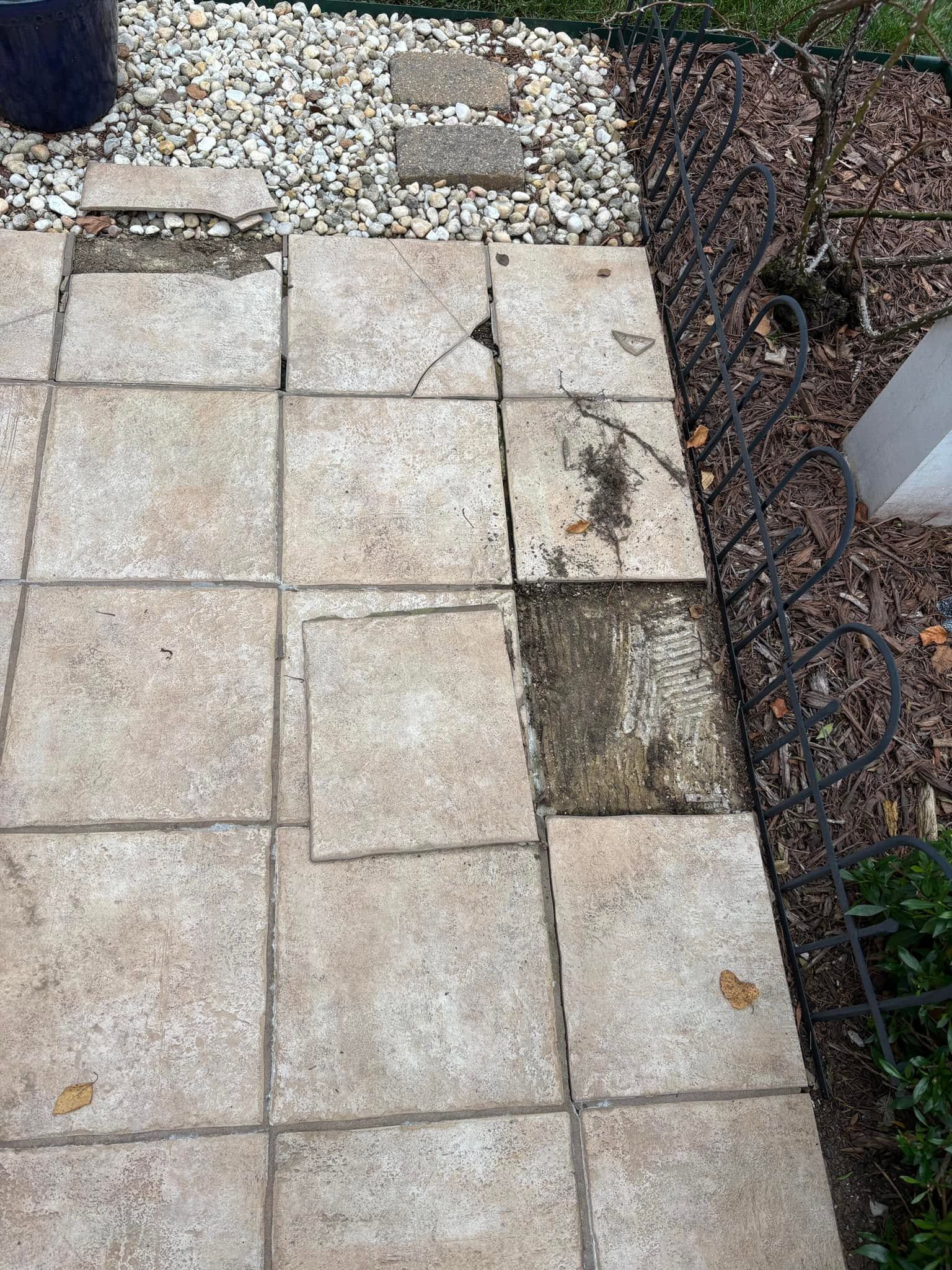 Damaged stone walkway with uneven cracked pavers beside gravel and a fence