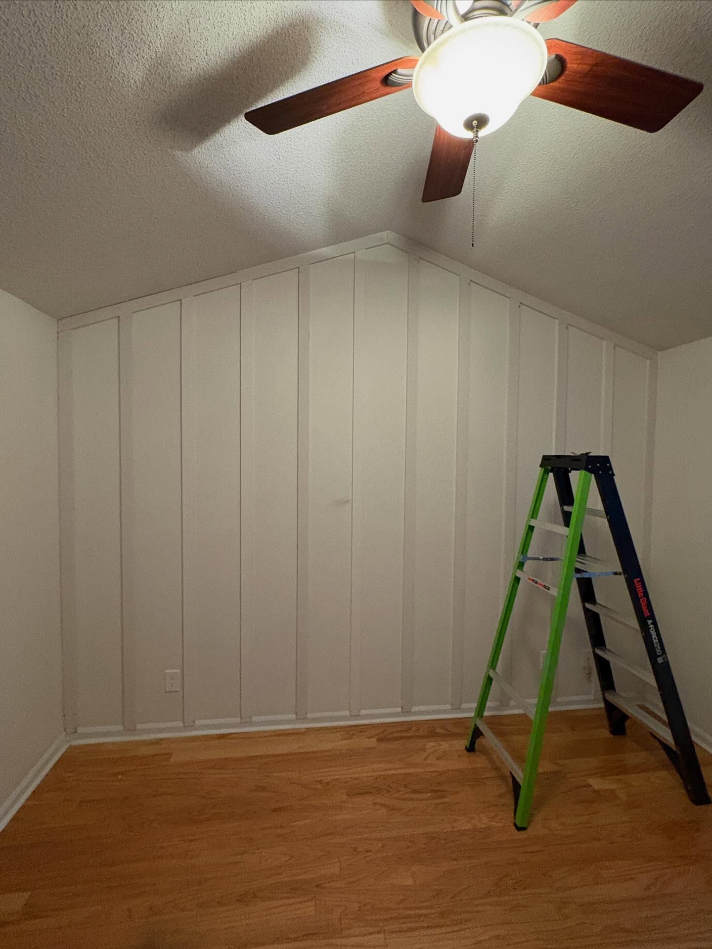Empty room with white paneled walls, wood floor, ceiling fan, and a green ladder.