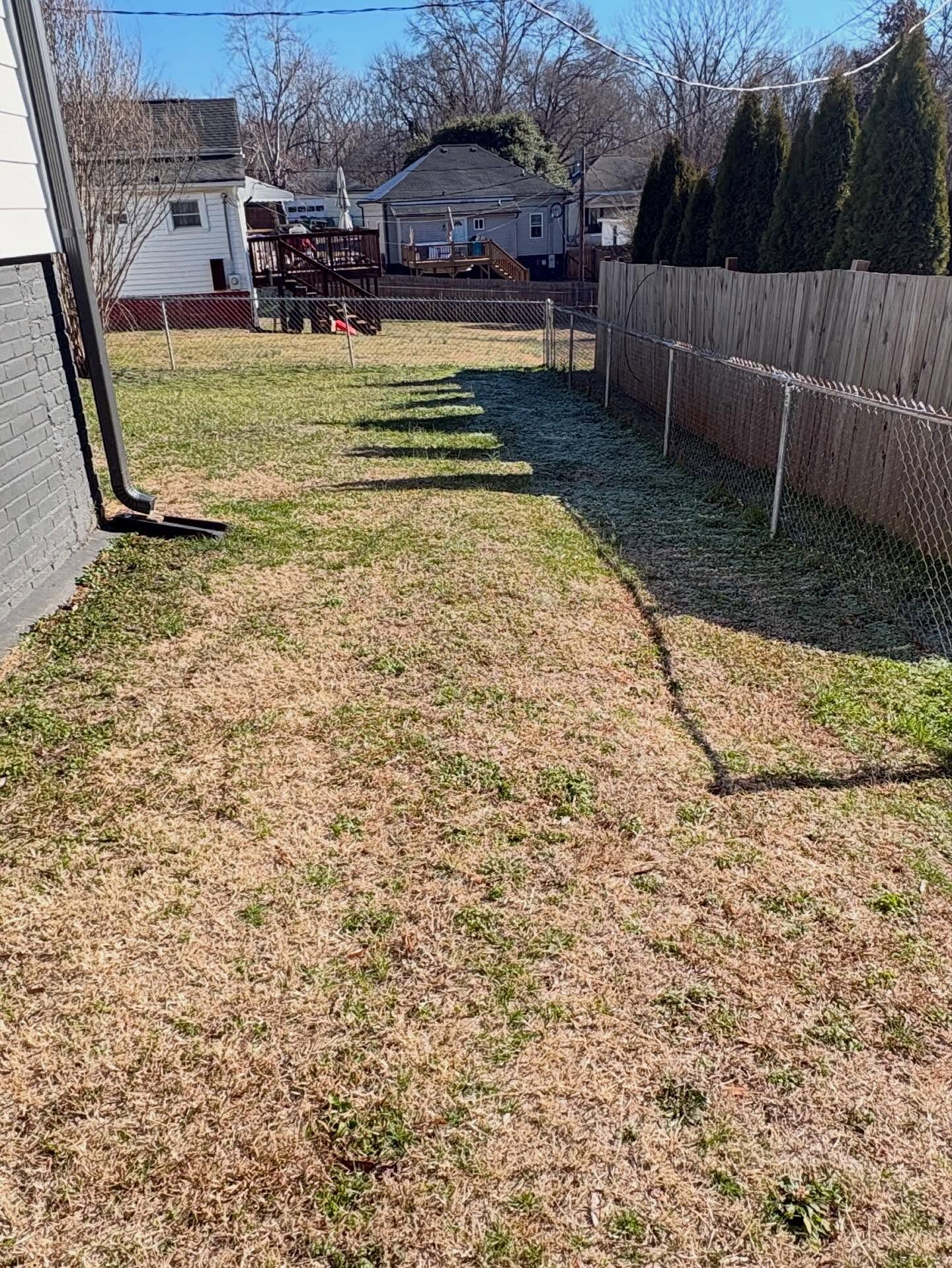 Narrow grassy backyard between a house and wooden fence, with leafless trees and a small deck at the far end