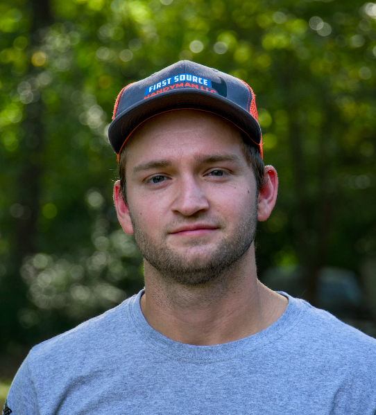Portrait of a man in a gray shirt and dark cap, standing outdoors with blurred green foliage behind him