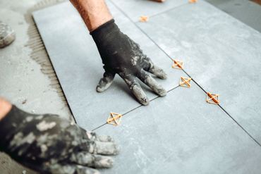 Gloved hands installing gray floor tiles with orange spacers on a tiled surface