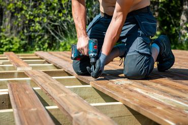 Person kneeling on a wooden deck frame, using a power drill to fasten planks outdoors.
