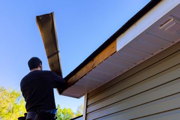 Person installing or repairing roof fascia board on a house exterior under blue sky