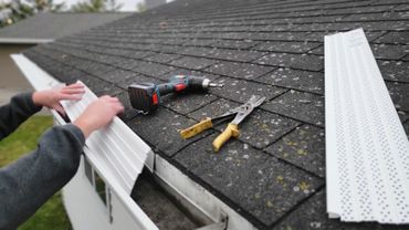 Person installing white gutter guards on a shingled roof with tools nearby