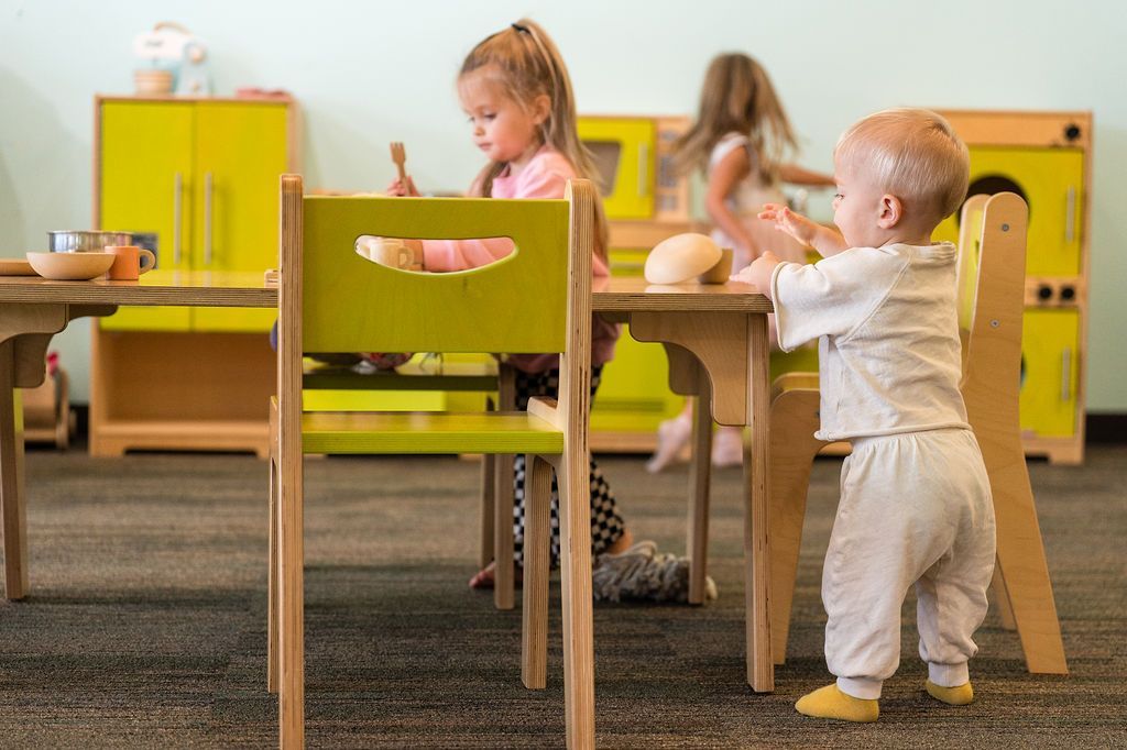 Children playing at small tables in a bright classroom with yellow chairs and toys