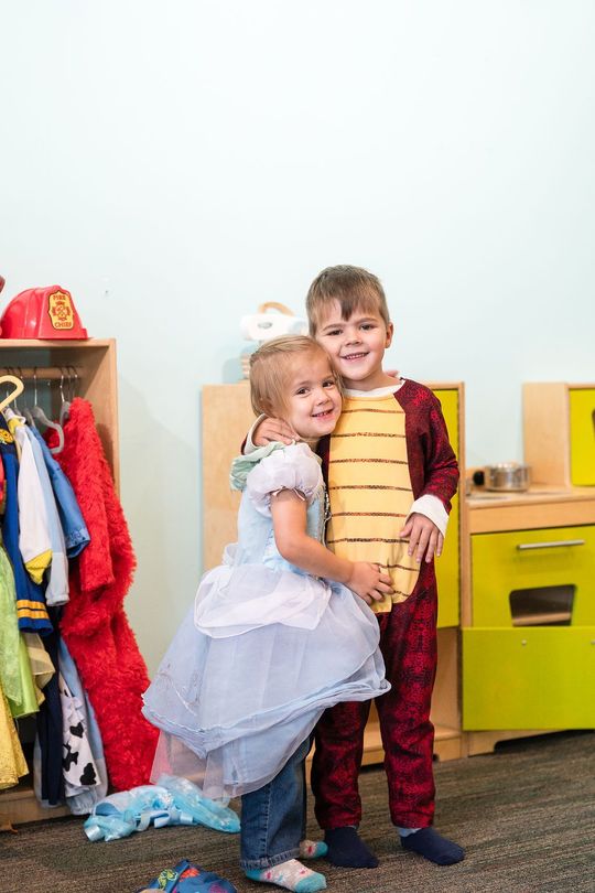 Two children hugging in a playroom, smiling beside colorful toy shelves.