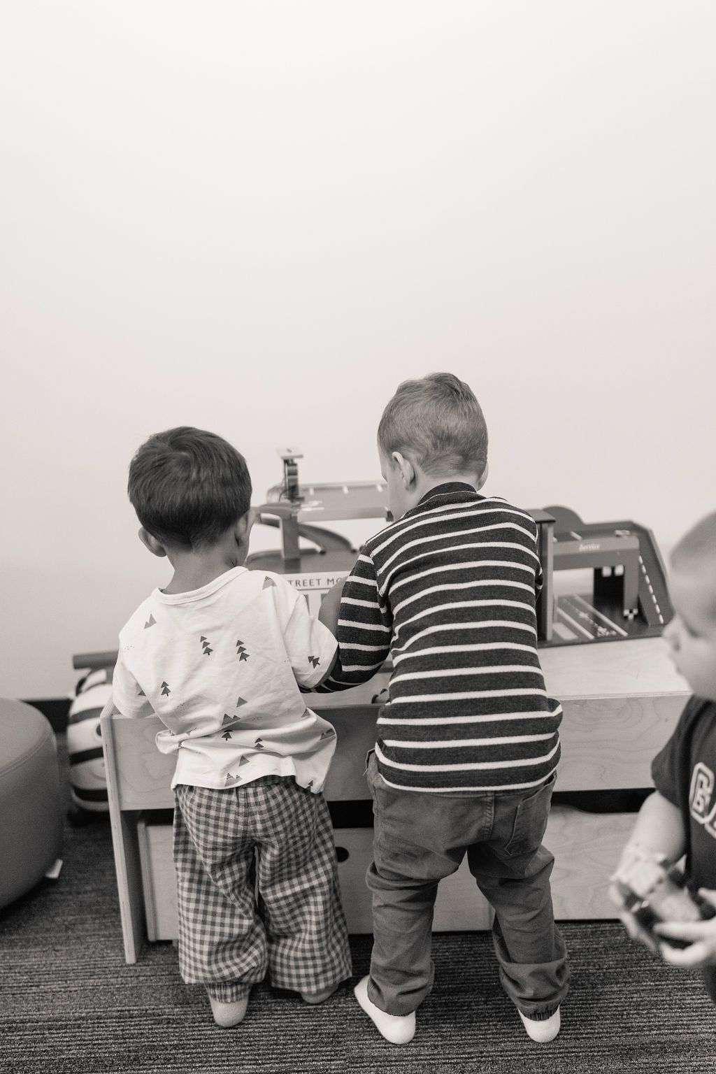 Two toddlers stand at a toy table, facing away, in a playroom.