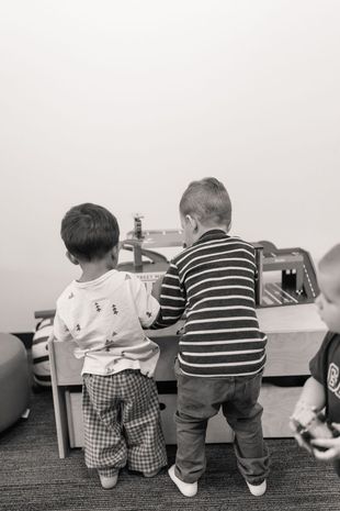 Two children standing at a toy train table, one in striped shirt and one in patterned clothes