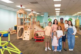 Group of people standing in a bright indoor playroom with wooden climbing structures and toys