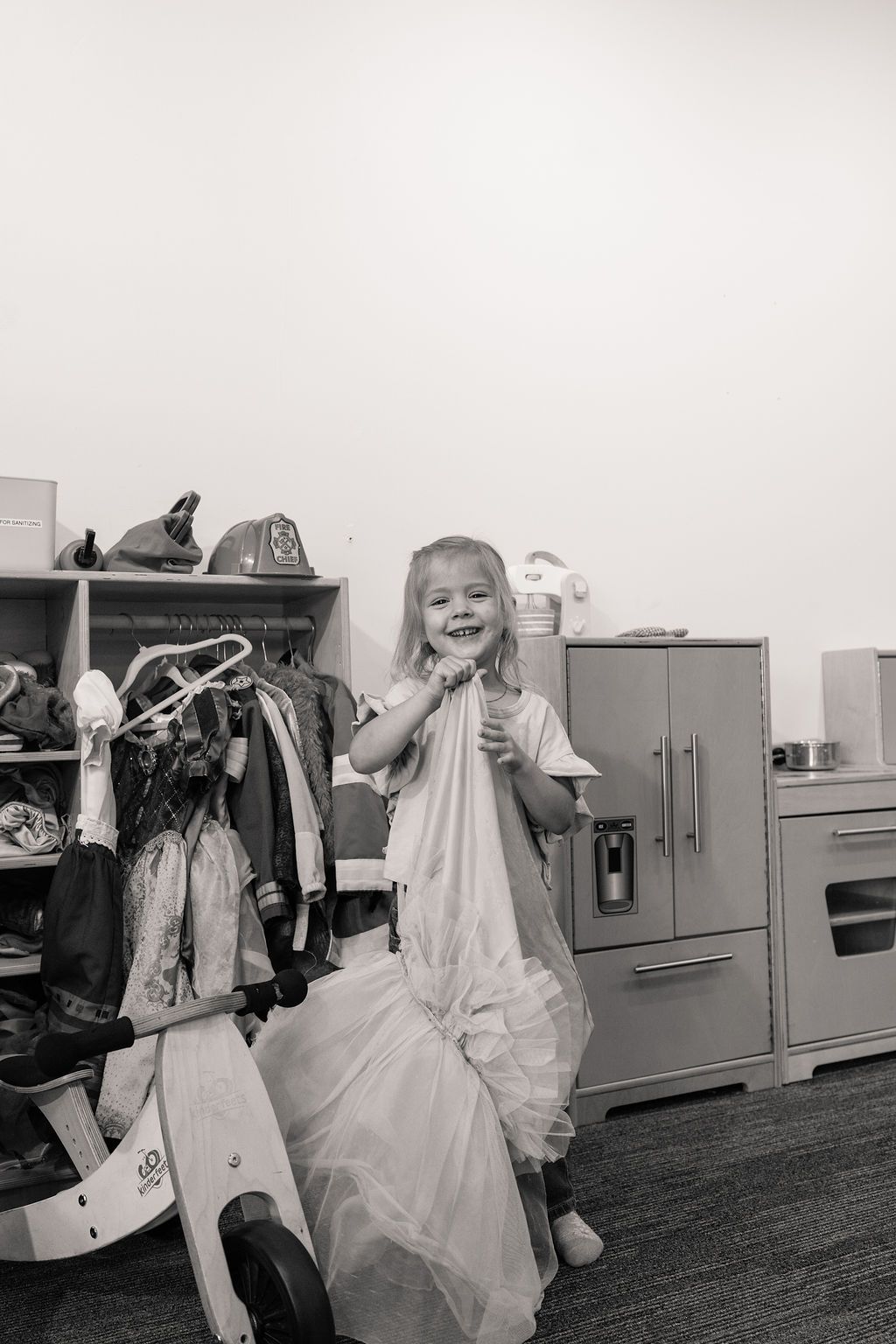 Child in a dress smiling and posing indoors beside a play kitchen and toy clothes rack.