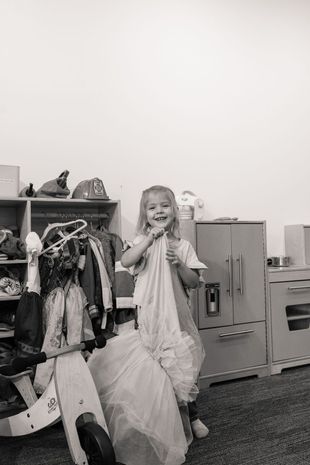 Child smiling and holding a toy microphone in a playroom beside a cabinet and hanging clothes
