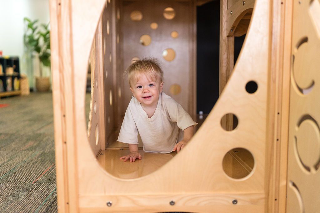 Child crawling through a wooden play structure, smiling in a daycare playroom.