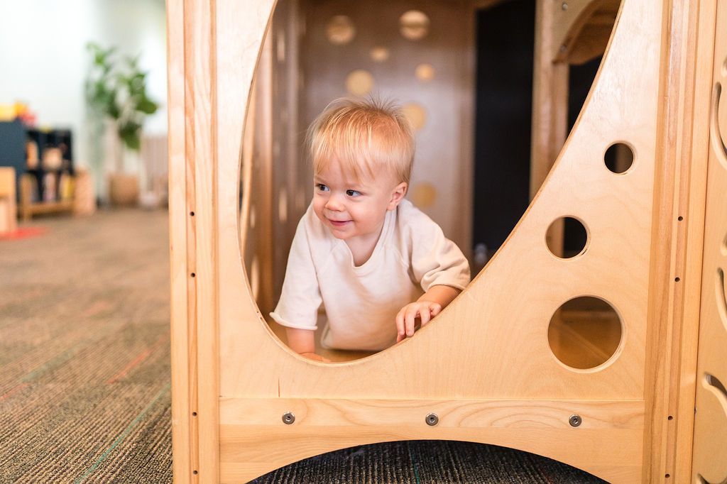 Toddler leaning through a wooden play structure, smiling in a bright indoor play area.