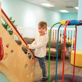 Child standing on indoor playground equipment, holding a tan climbing wall with colorful bars nearby.