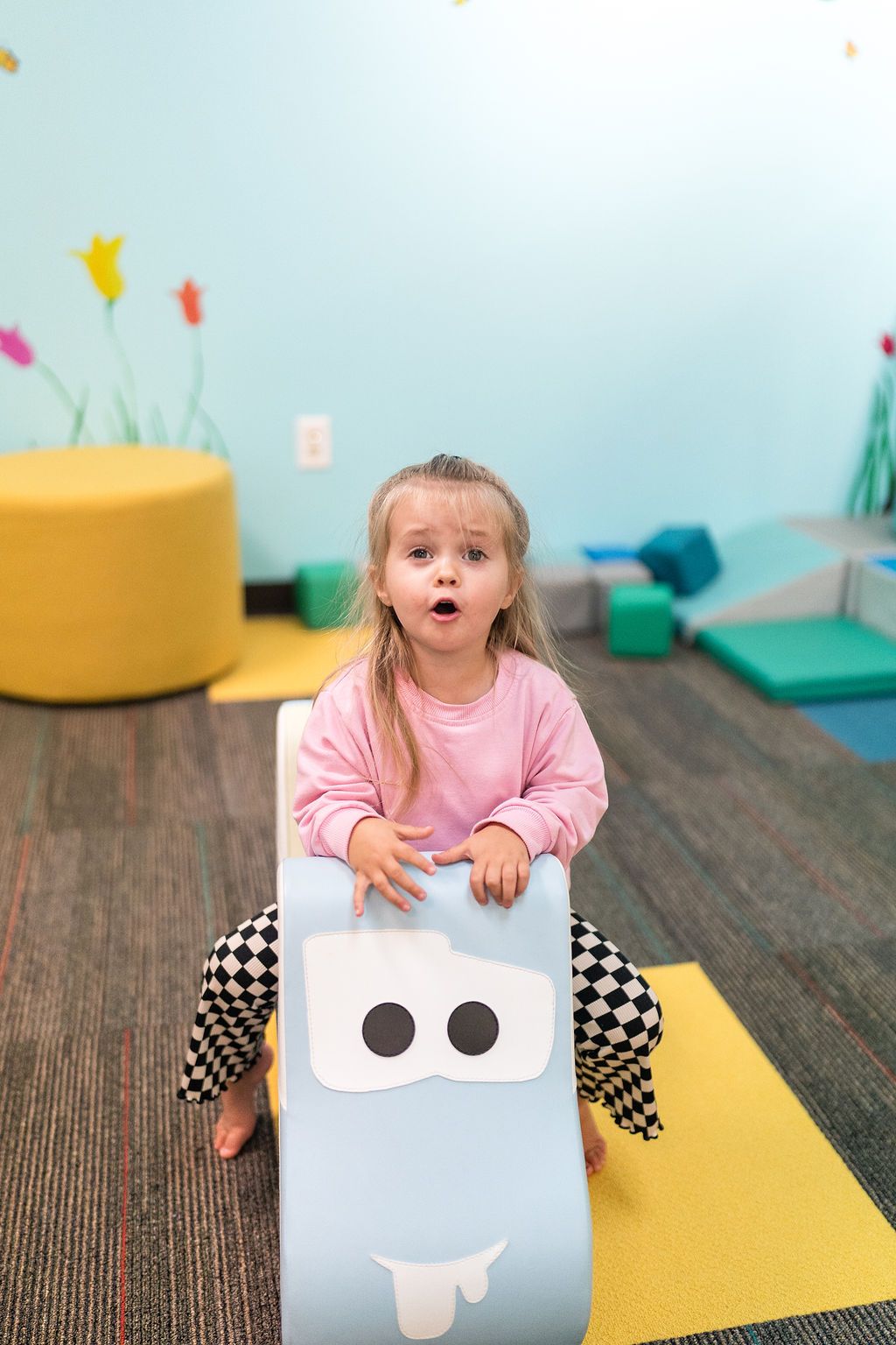 Child in a pink shirt sitting on a toy in a playroom, looking surprised.