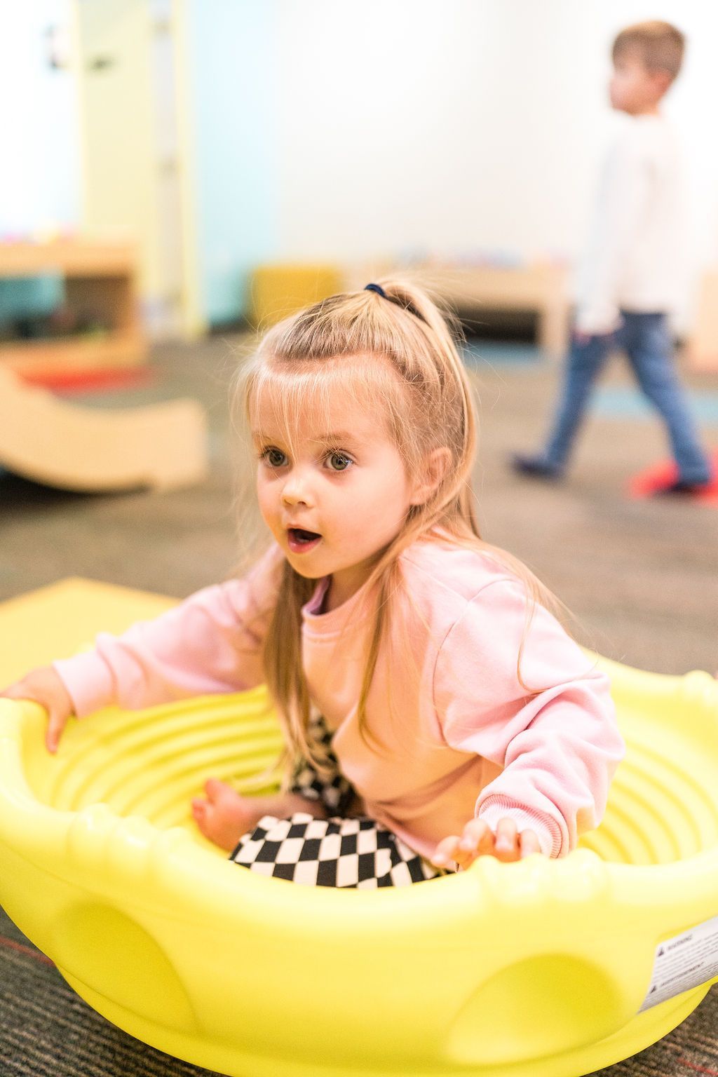 Child in a yellow inflatable ring at an indoor play area, looking surprised with a checkered toy in front.