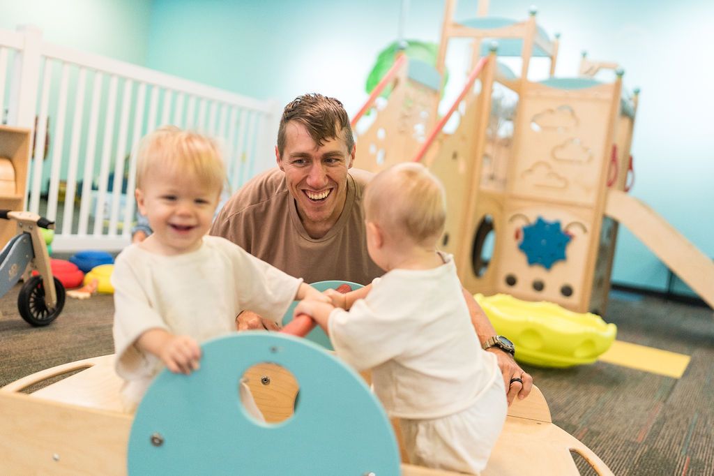 Adult and two children playing on colorful indoor playground equipment, smiling on a padded floor.