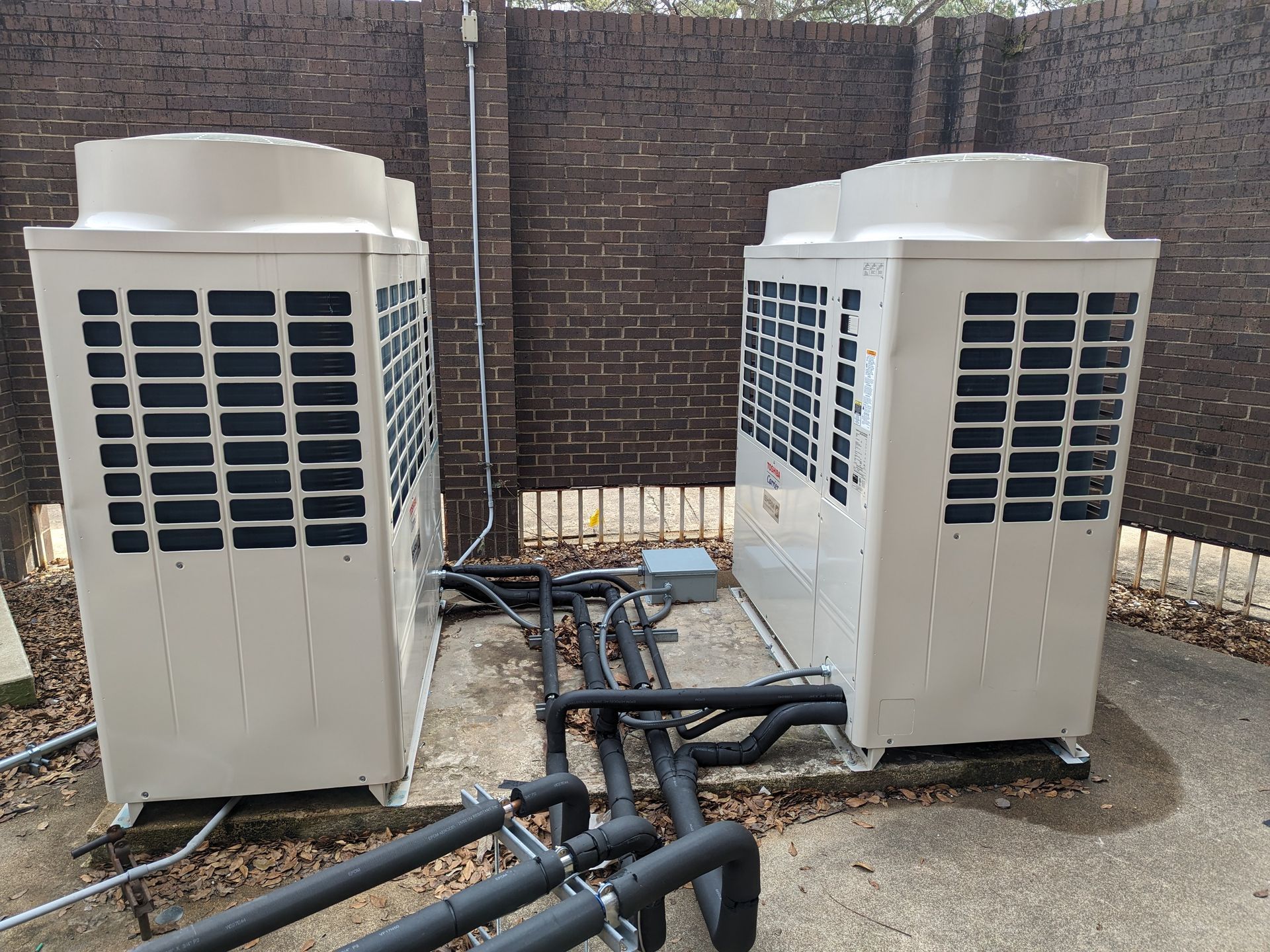 Two air conditioners are sitting next to each other in front of a brick wall.