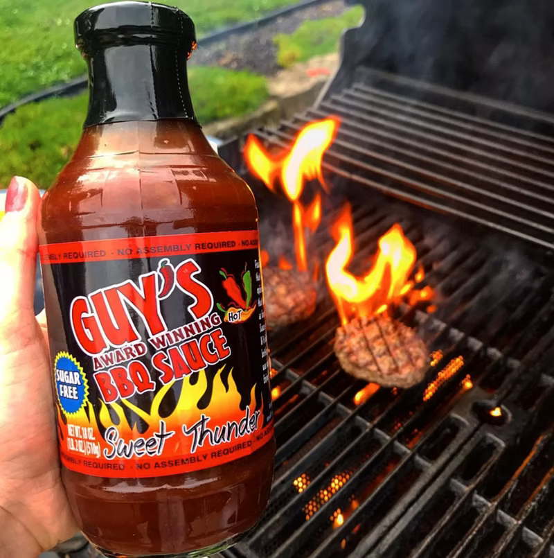 Guns Sauce barbecue sauce bottle beside a grill with meat flames and smoke indoors