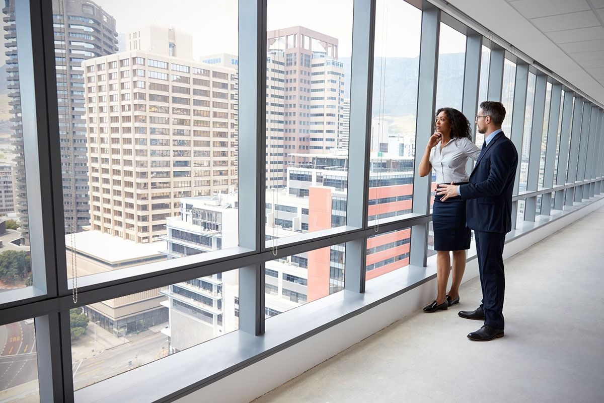 Two business people stand by a windowed office corridor overlooking city buildings.