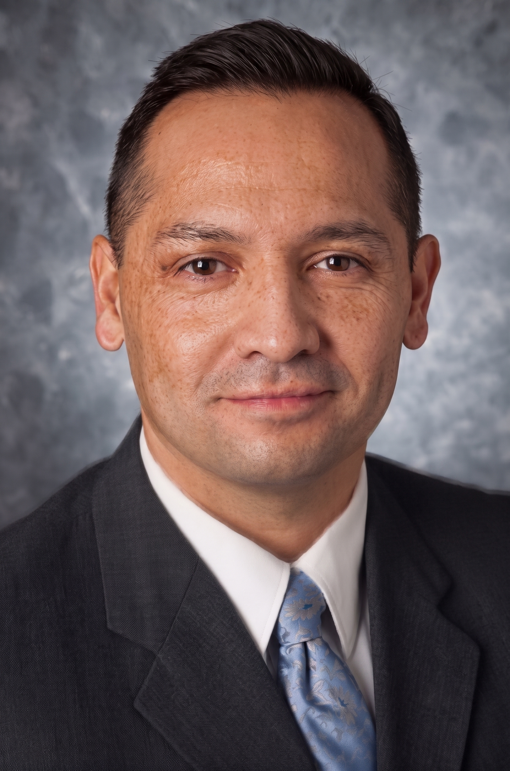 Studio headshot of a man in a dark suit and blue tie against a gray backdrop