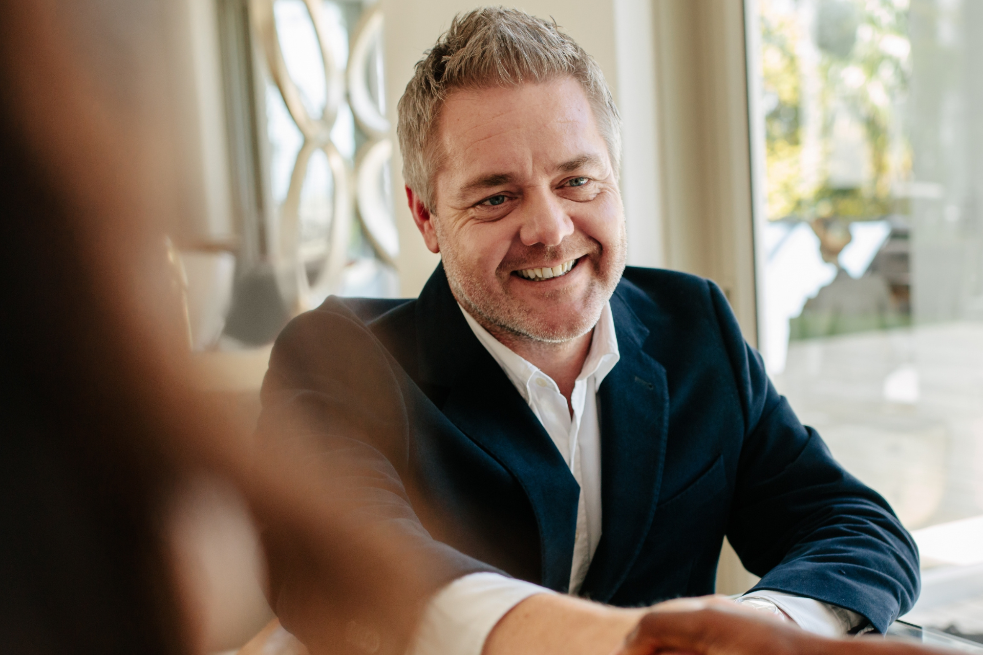 Smiling man in a dark blazer seated at a table in a bright room, talking with someone out of focus.