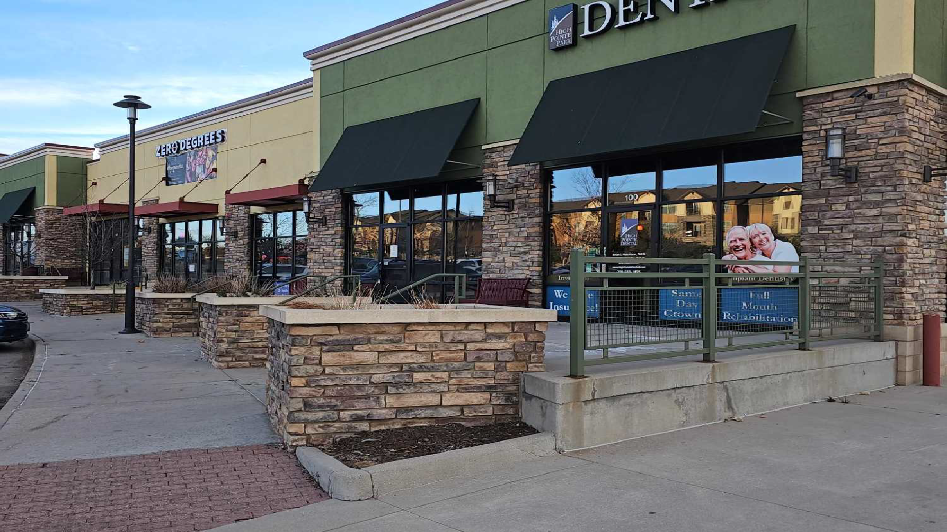 Strip mall storefronts with green awnings, stone planters, and a sidewalk in front.