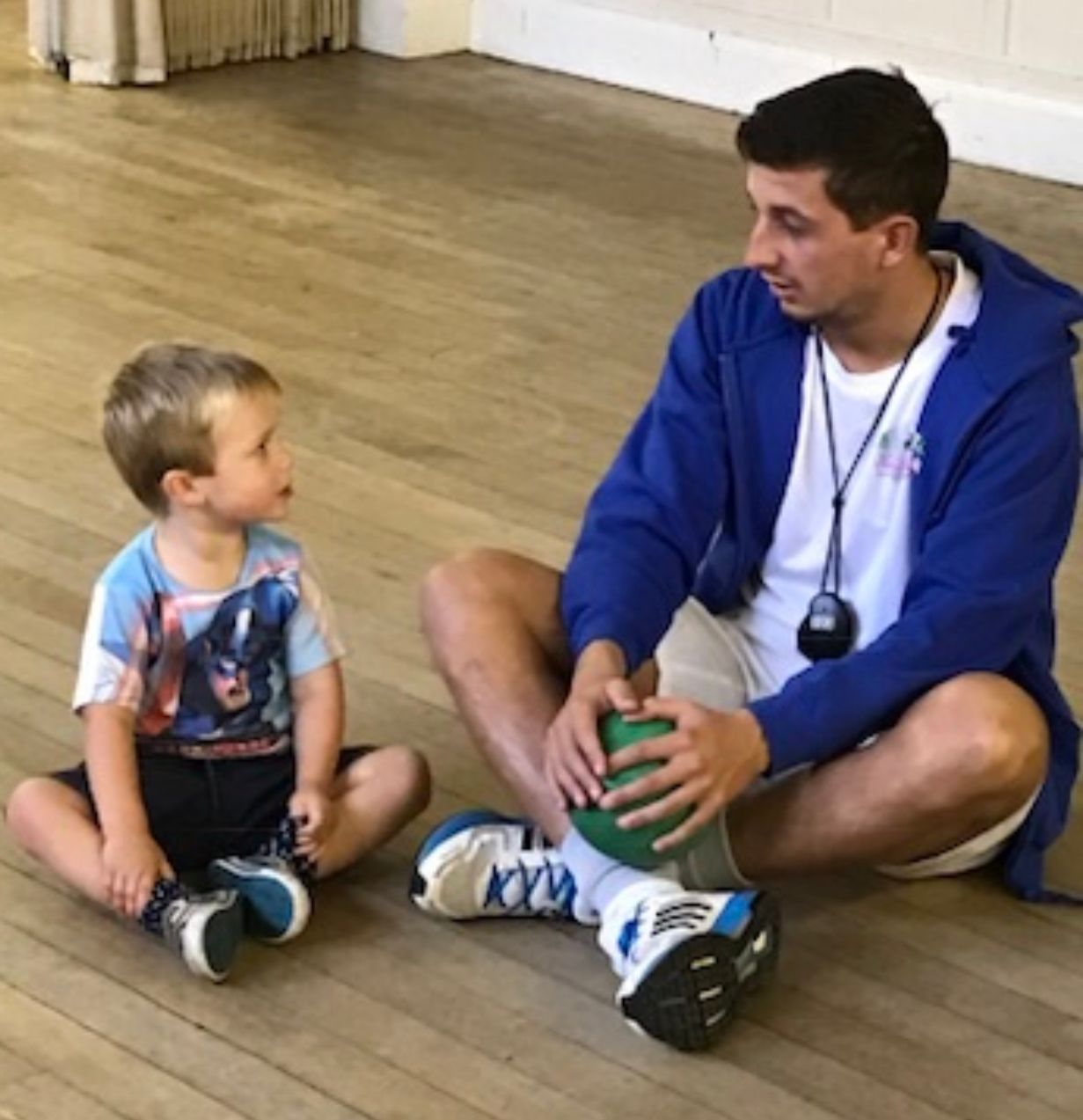 A man in a blue jacket sits on the floor talking to a little boy