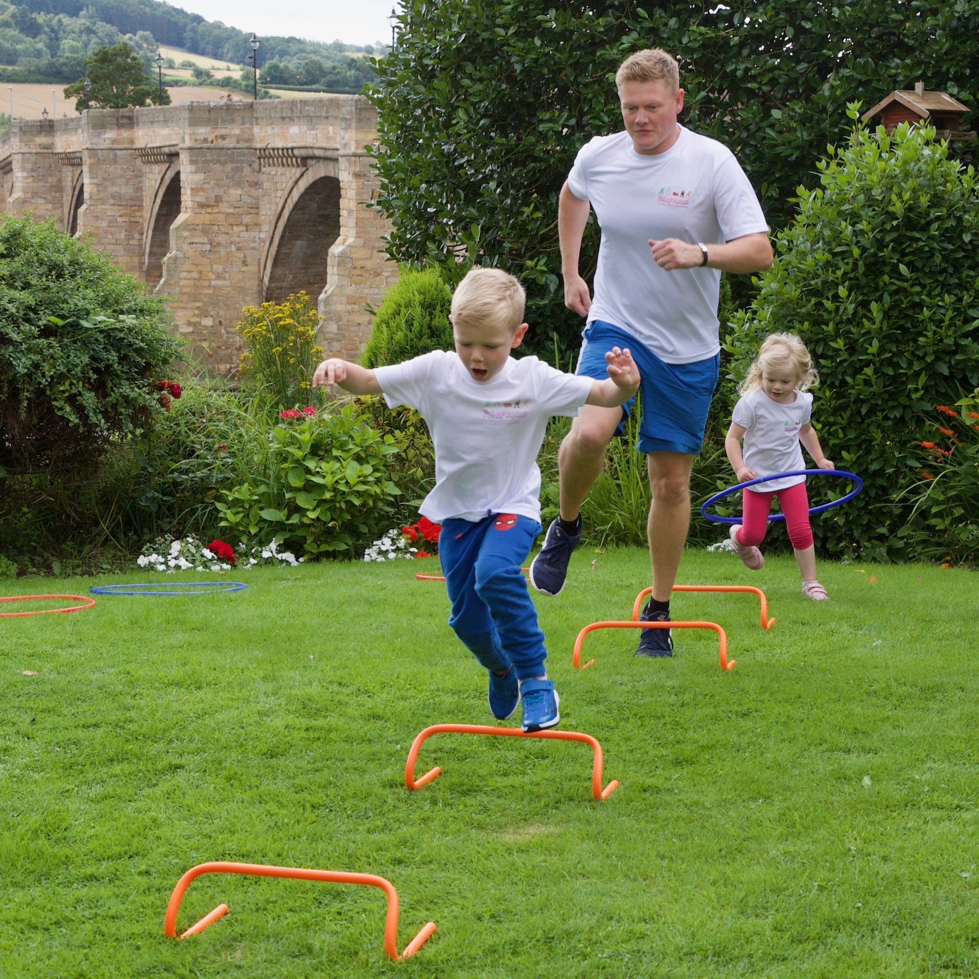 A man and two children are jumping over hurdles on a lush green field.