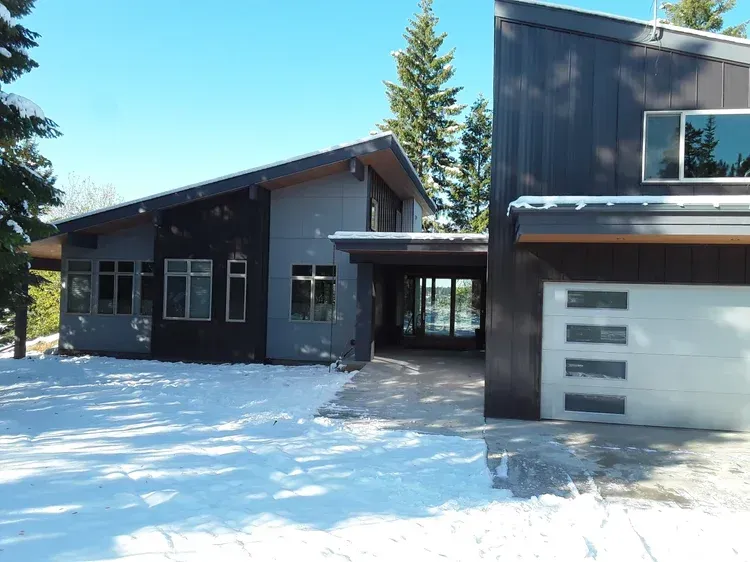 Snow-covered modern house exterior with dark siding, garage, and tall evergreen trees.