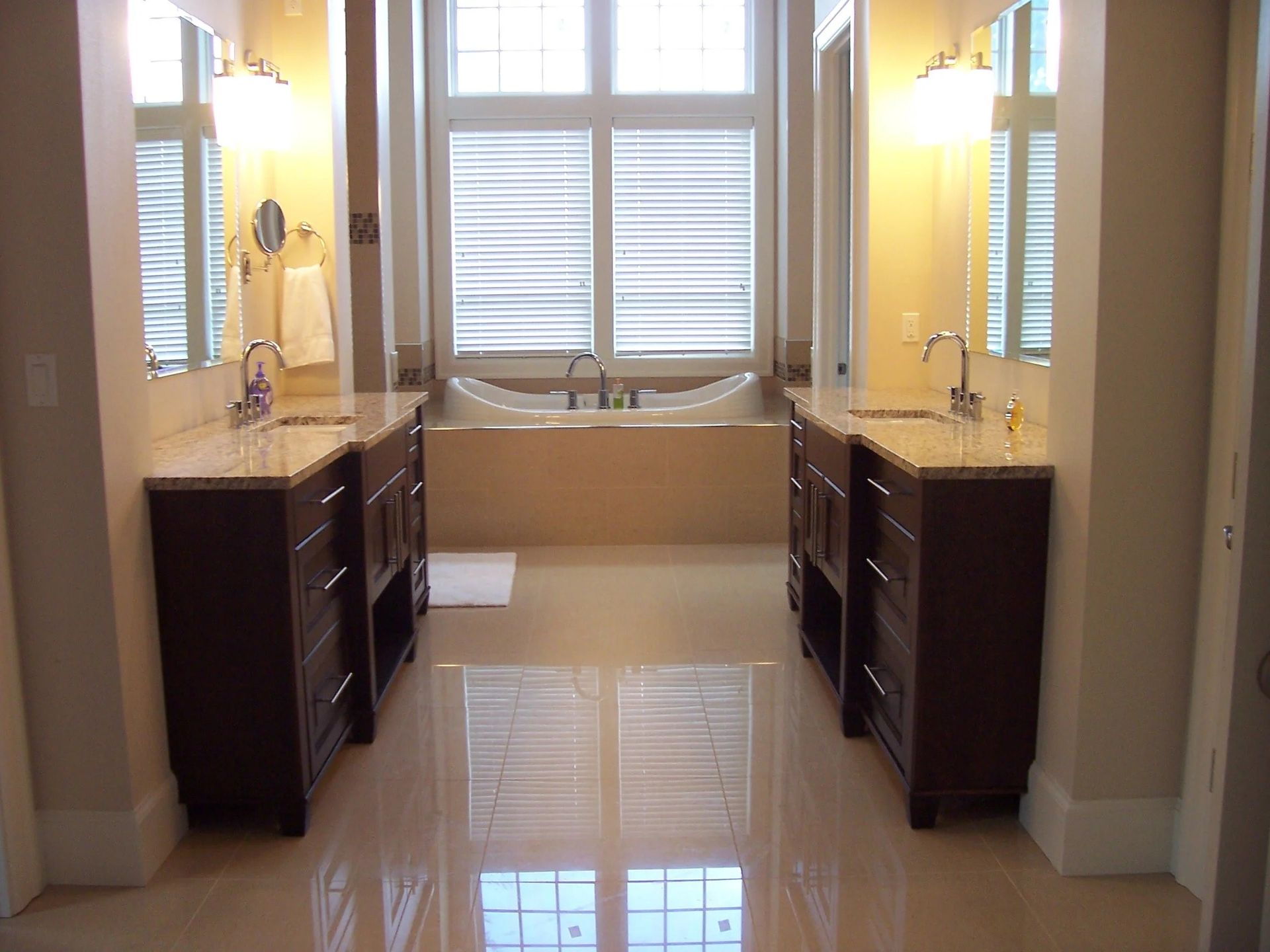 Bright bathroom with twin sinks, dark vanities, glossy tile floor, and a window with white blinds.