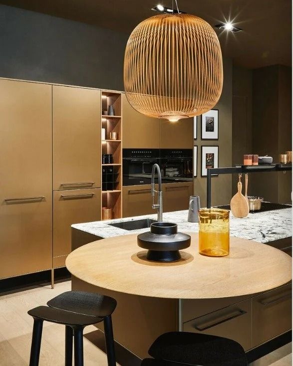Modern kitchen with a round wooden island, black stool, gold cabinets, and woven pendant light.