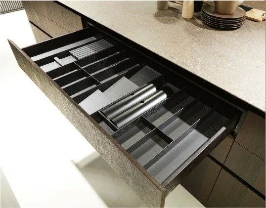 Kitchen drawer with adjustable black dividers and stacked silver utensils on a countertop.