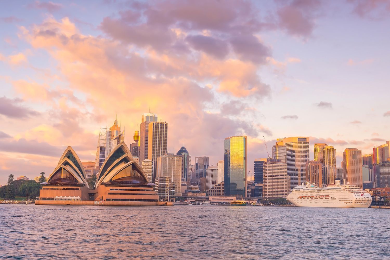 Sydney Opera House and skyline at sunset across the harbor with glowing clouds and a ferry on the water