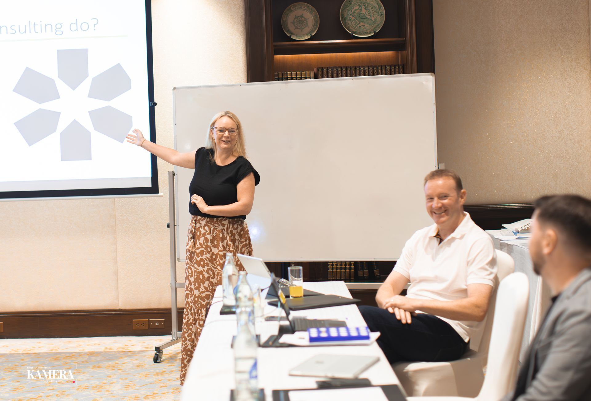 Presenter pointing at a projection screen during a meeting, with attendees seated at a table and a whiteboard nearby.