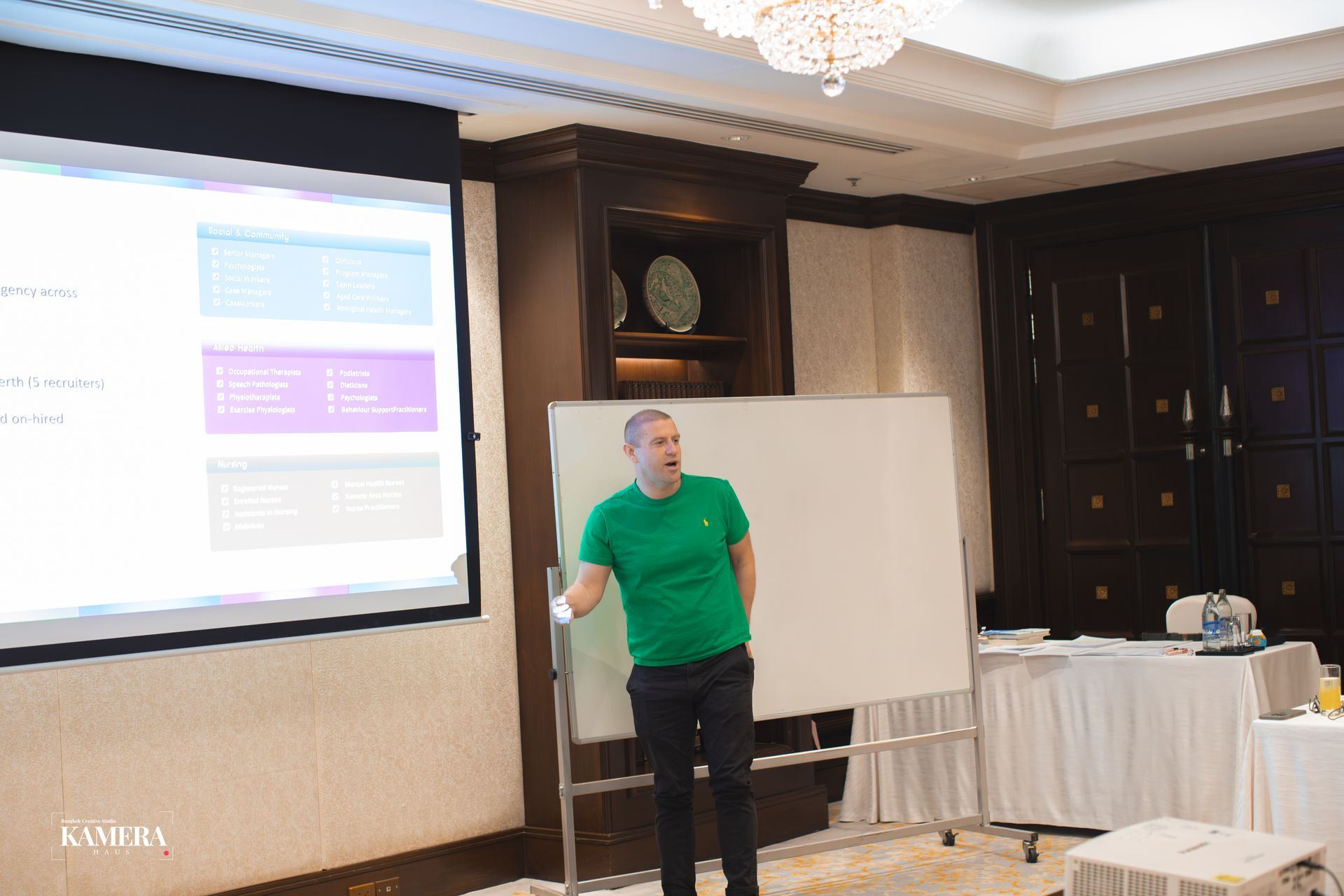 Presenter in a green shirt speaking beside a projection screen in a conference room