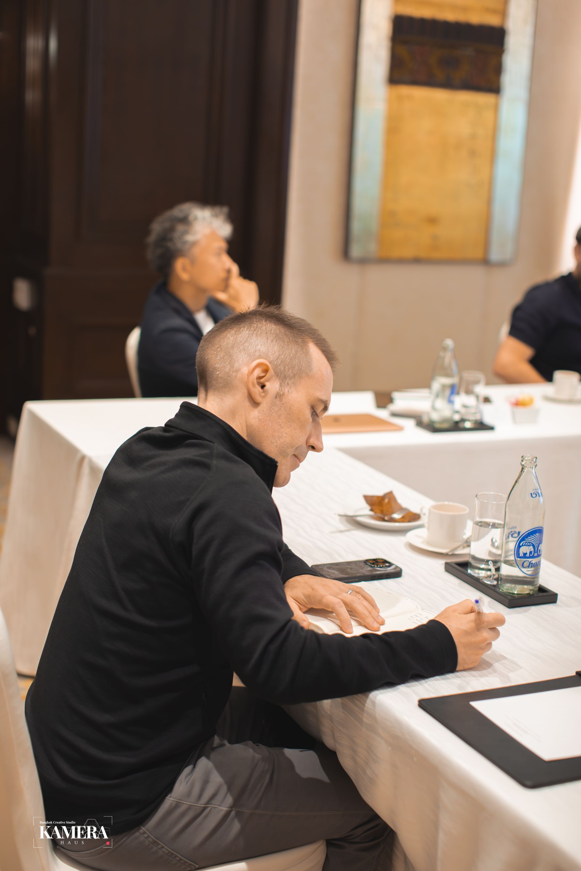 Person seated at a conference table, writing notes in a meeting room with cups and water bottles.