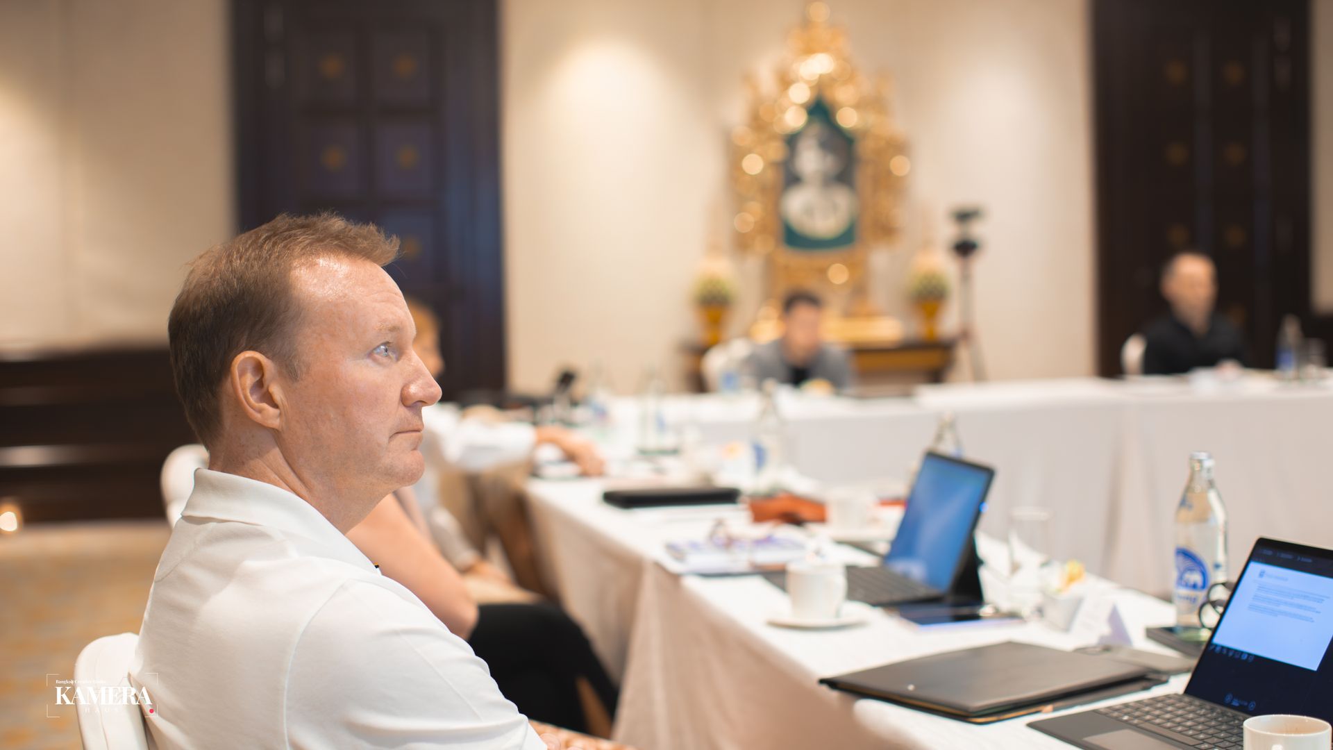 Man seated at a conference table with laptops and papers in a banquet hall, looking toward the front.