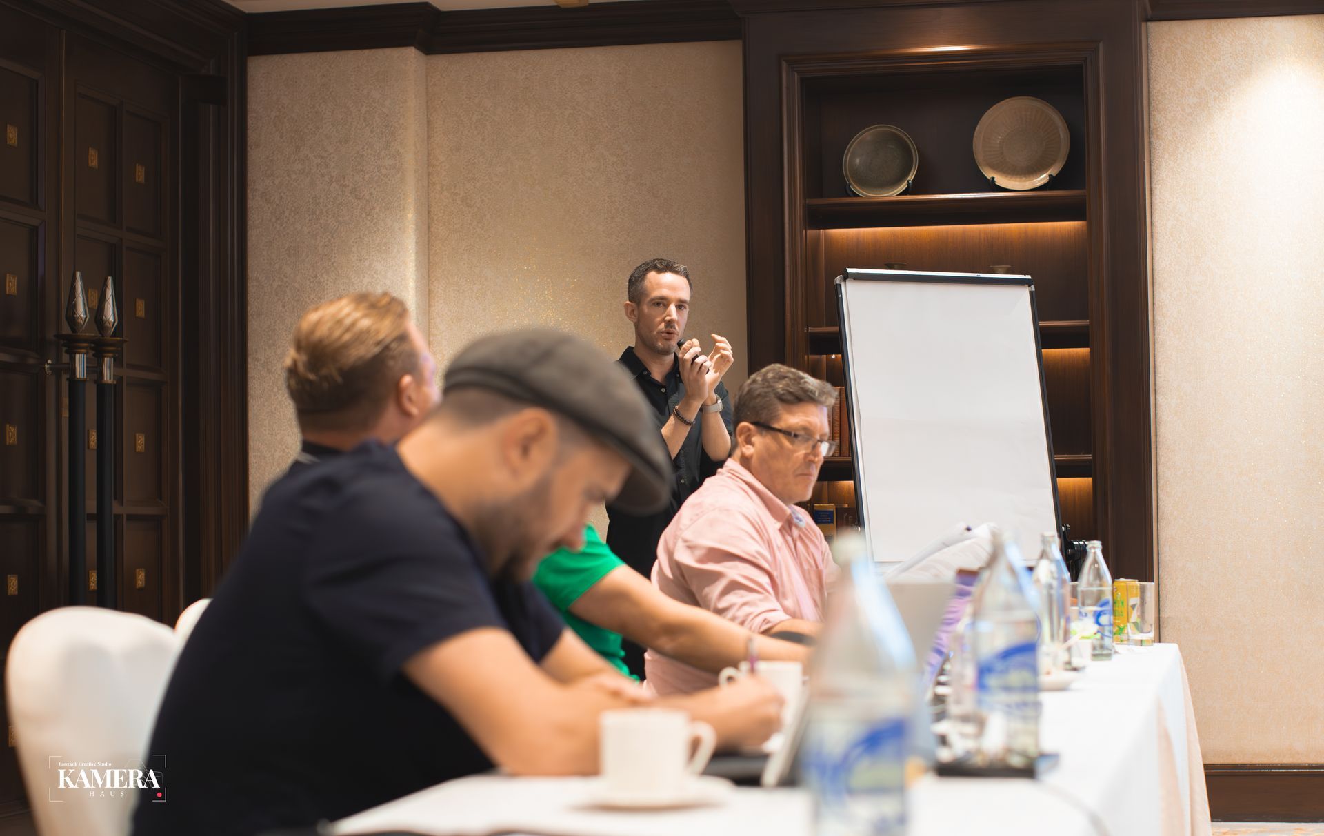 People seated at a conference table during a meeting, with a presenter standing by a flip chart.