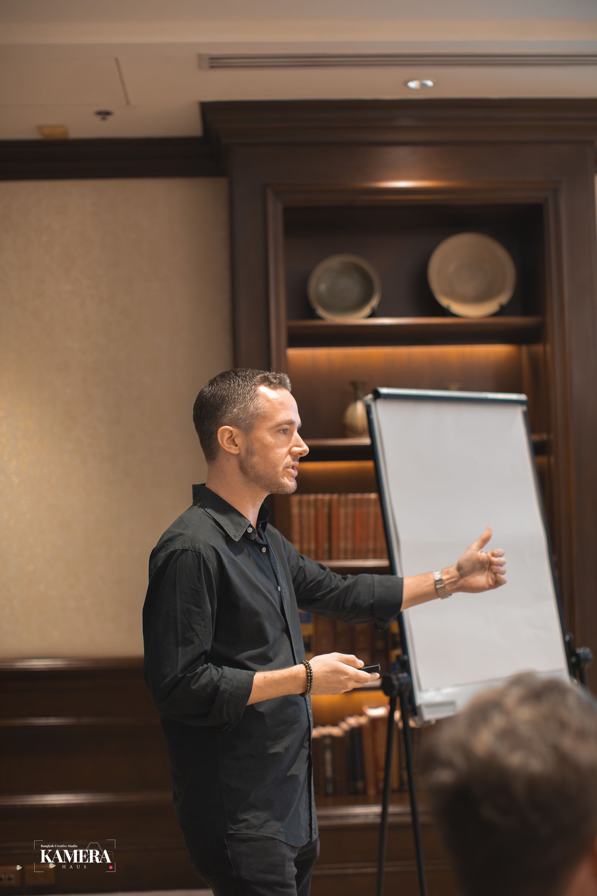 Man presenting beside a flip chart in a conference room, gesturing toward the board