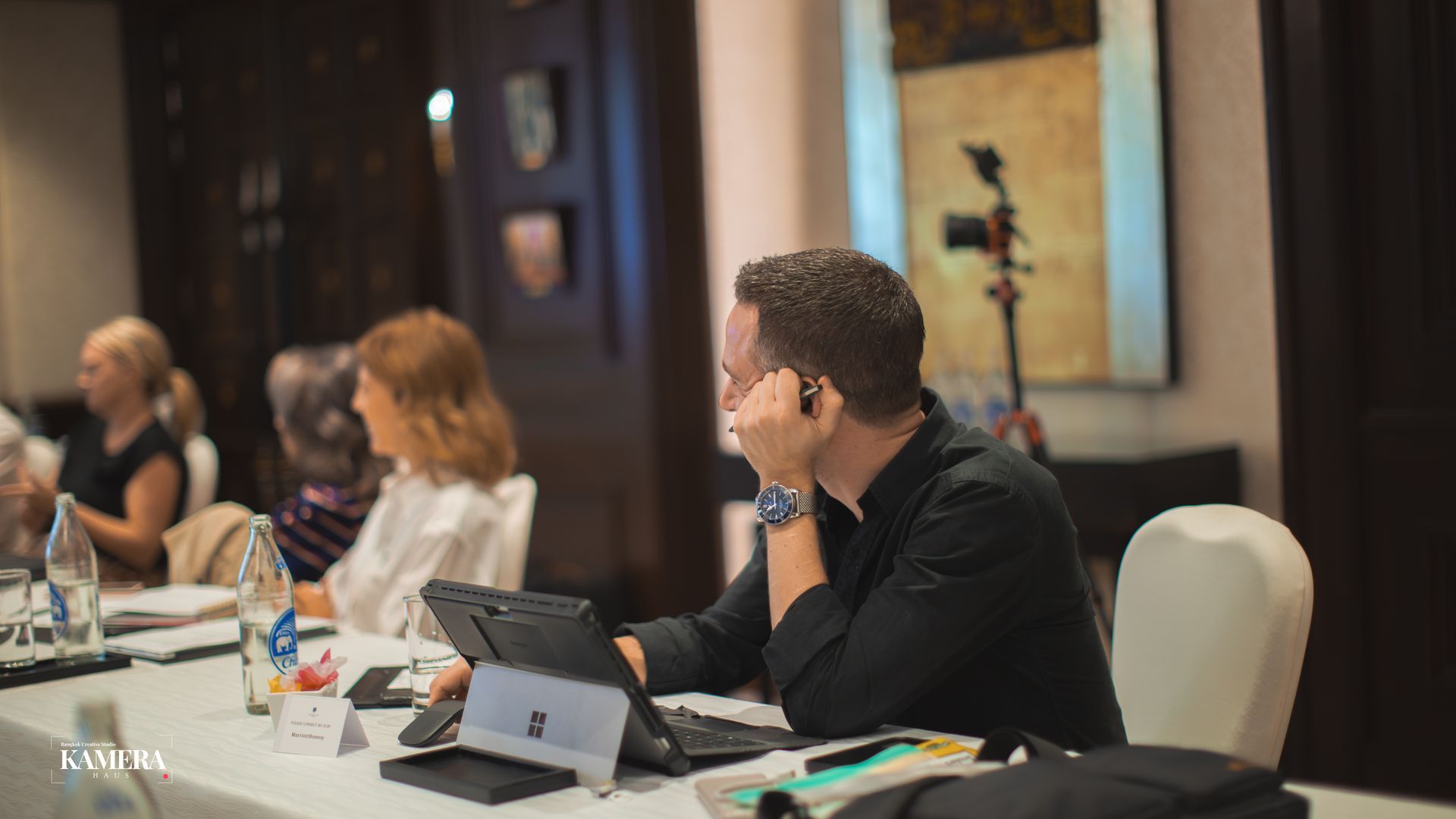 Conference attendees seated at a table, one man using a tablet and microphone in the foreground.