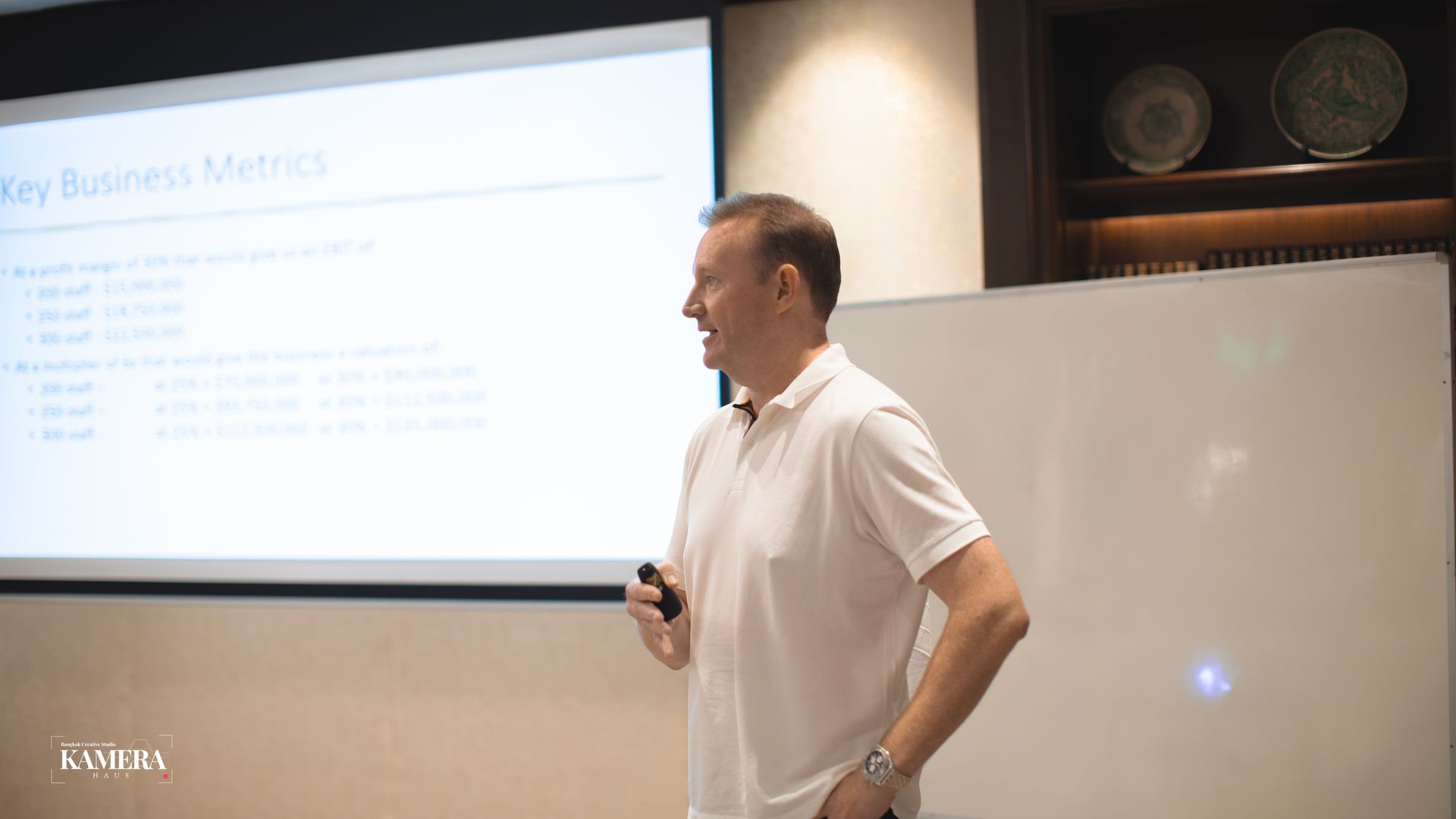 Man presenting beside a projected slide in a conference room, holding a remote.