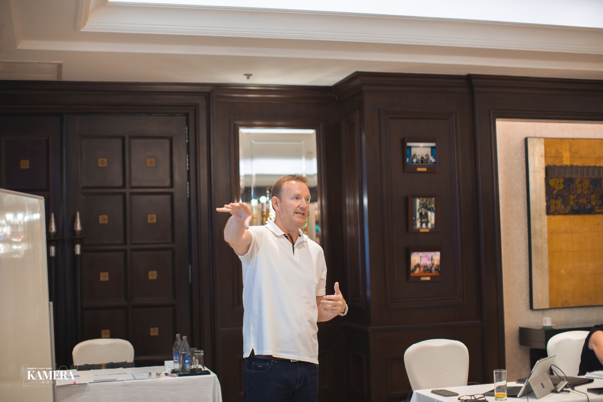 Man speaking and gesturing in a conference room with dark wood paneling and tables set up