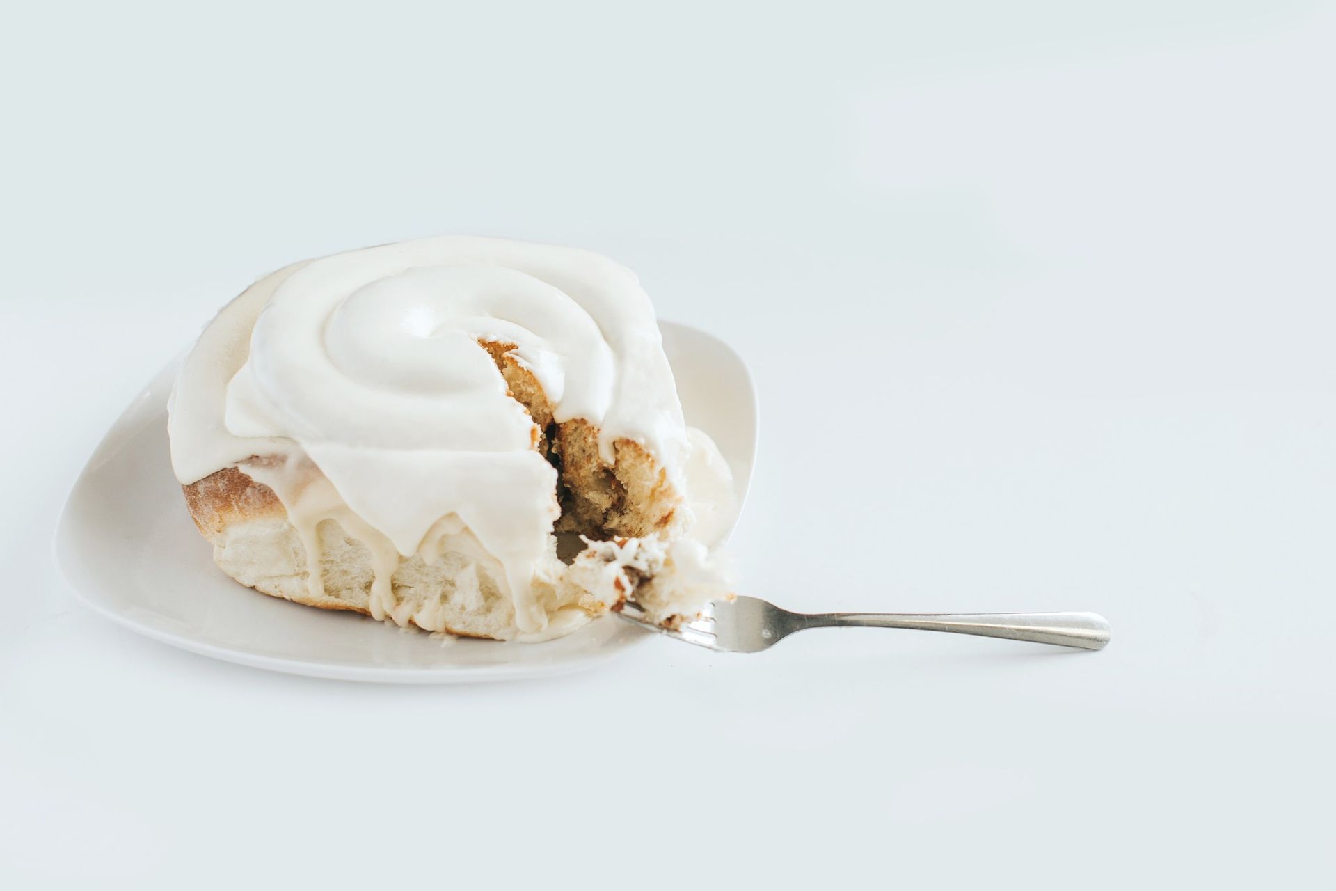 A cinnamon roll topped with white icing on a white plate, with a fork beside it.